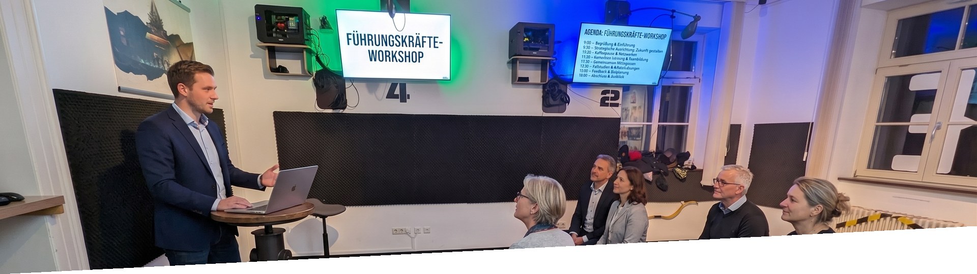 Führungsteam im Workshop: Berater am Stehpult, Teilnehmende im Halbkreis, Bildschirm mit dem Titel Führungskräfte-Workshop